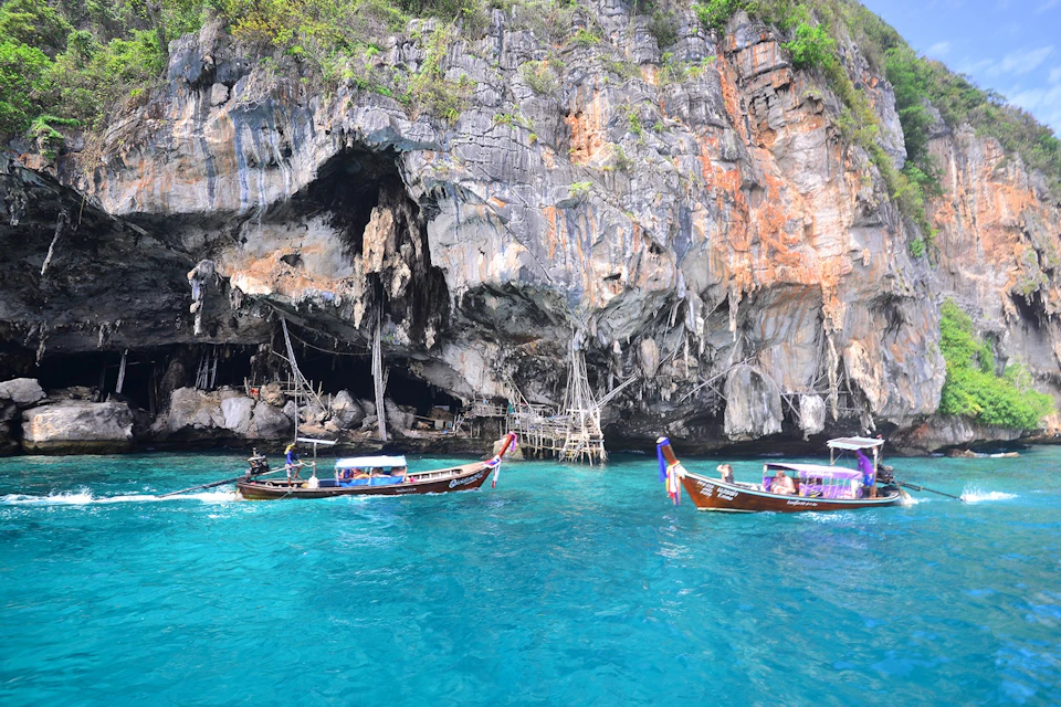 Two longtail boats float on clear turquoise water near a rocky cliff with cave openings and wooden structures.