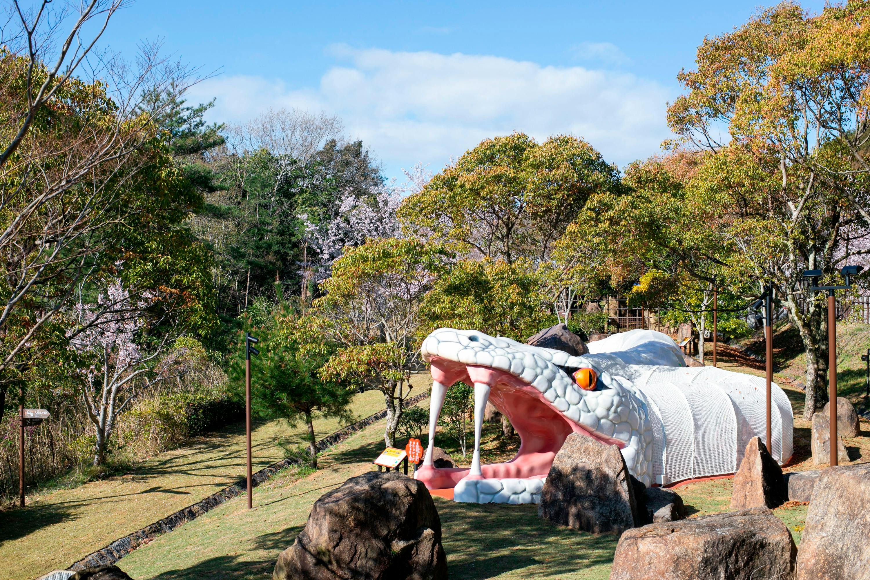 A large sculpture of a white snake with fangs in a park setting, surrounded by trees and rocks under a blue sky.