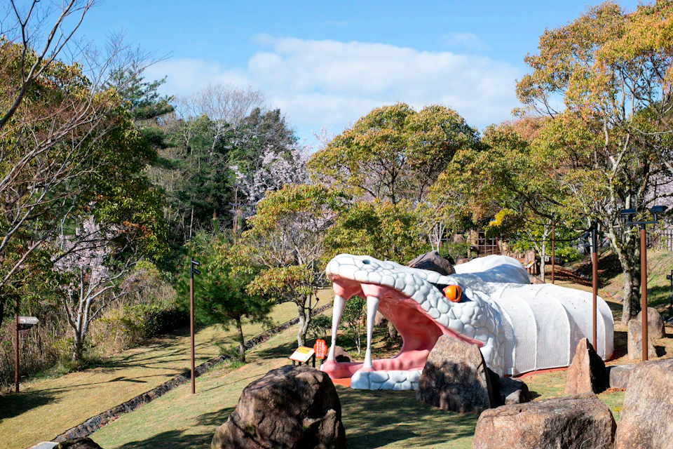 A large snake sculpture with an open mouth is set in a park surrounded by grass, trees, and rocks under a blue sky.