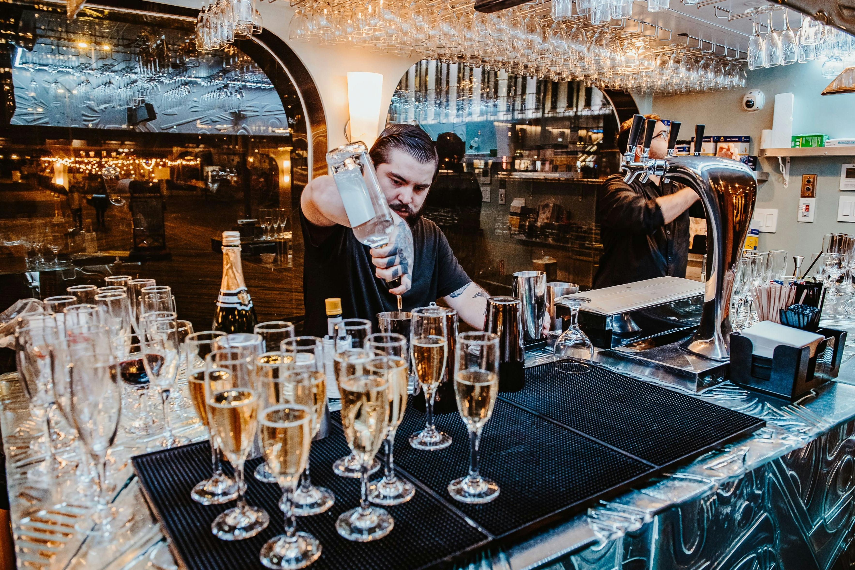 Barman nalévá nápoje do sklenic na šampaňské v baru s reflexním pozadím a závěsným skleněným nádobím.