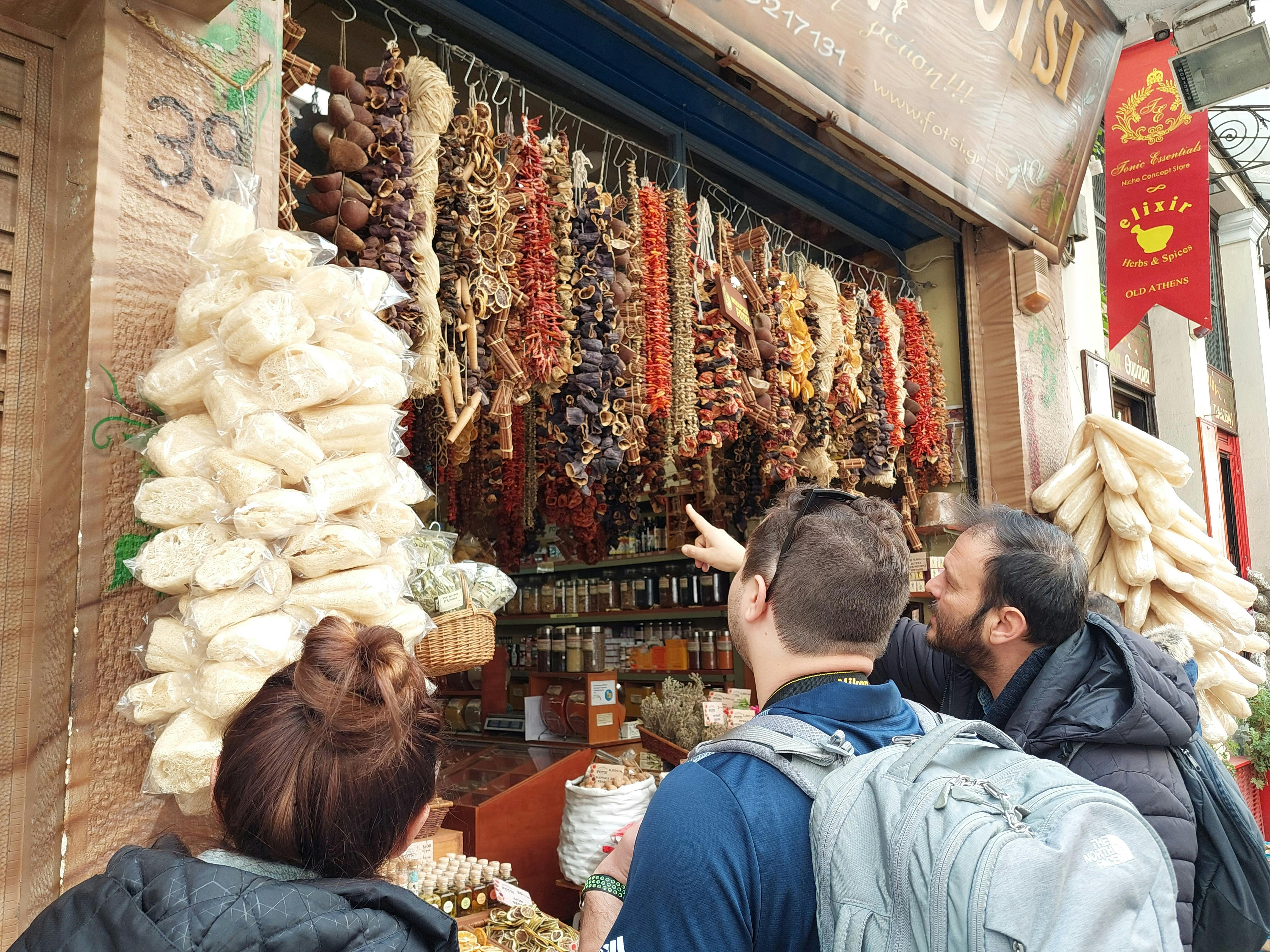 Τρεις άνθρωποι στέκονται μπροστά από έναν πάγκο γεμάτο με κρεμασμένα αποξηραμένα τρόφιμα και βάζα, με ένα άτομο να δείχνει προς τα αντικείμενα.