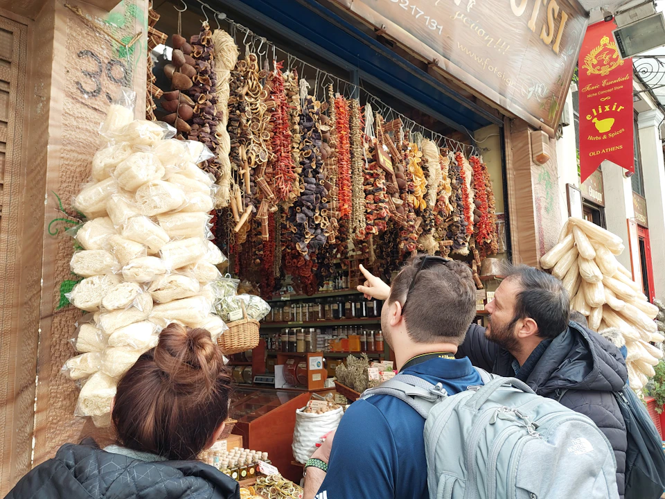 Three people stand in front of a stall filled with hanging dried foods and jars, with one person pointing towards the items.