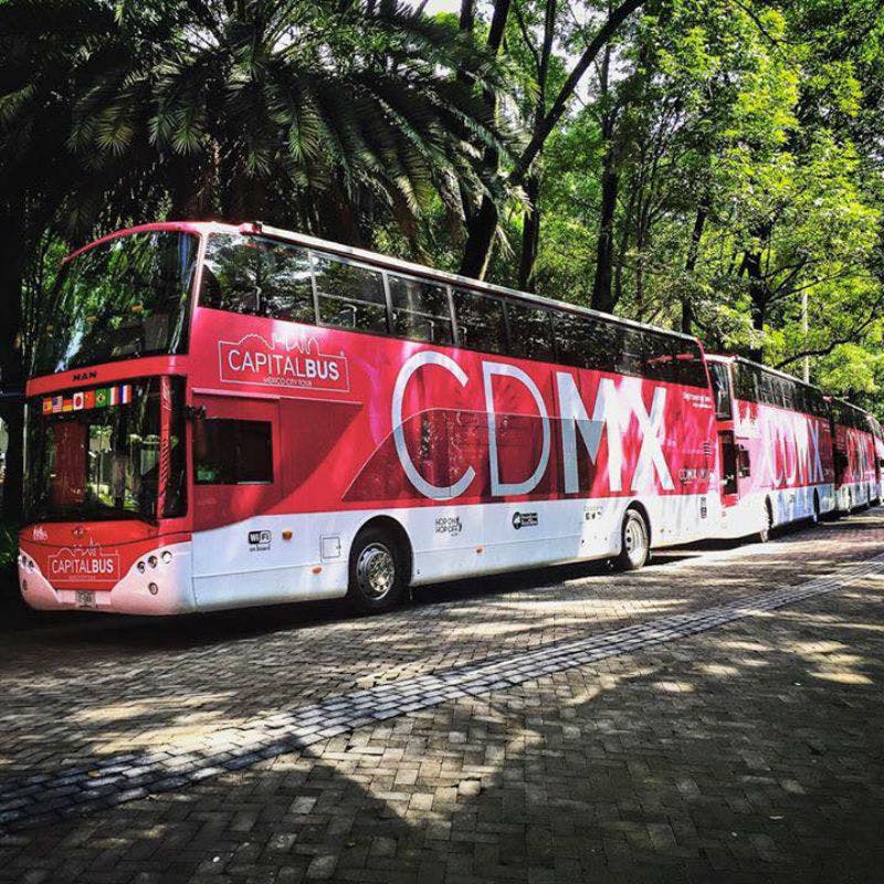 Zářivě červený dvoupatrový zájezdový autobus s nápisy „CDMX“ a „Capitalbus“ zaparkovaný na ulici lemované stromy.