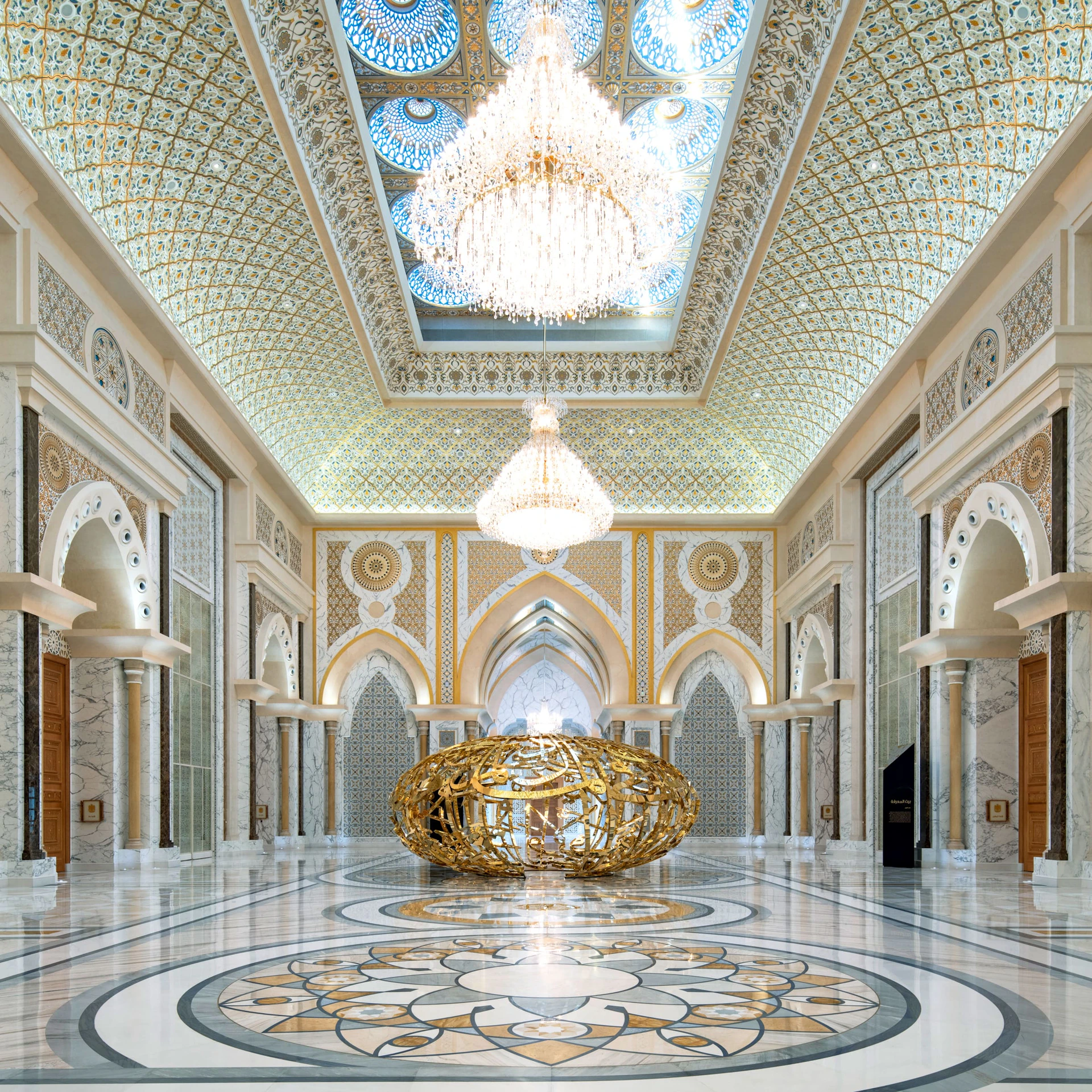 A grand hall with ornate arches, chandeliers, intricate ceiling designs, and a large gold sculpture in the center.