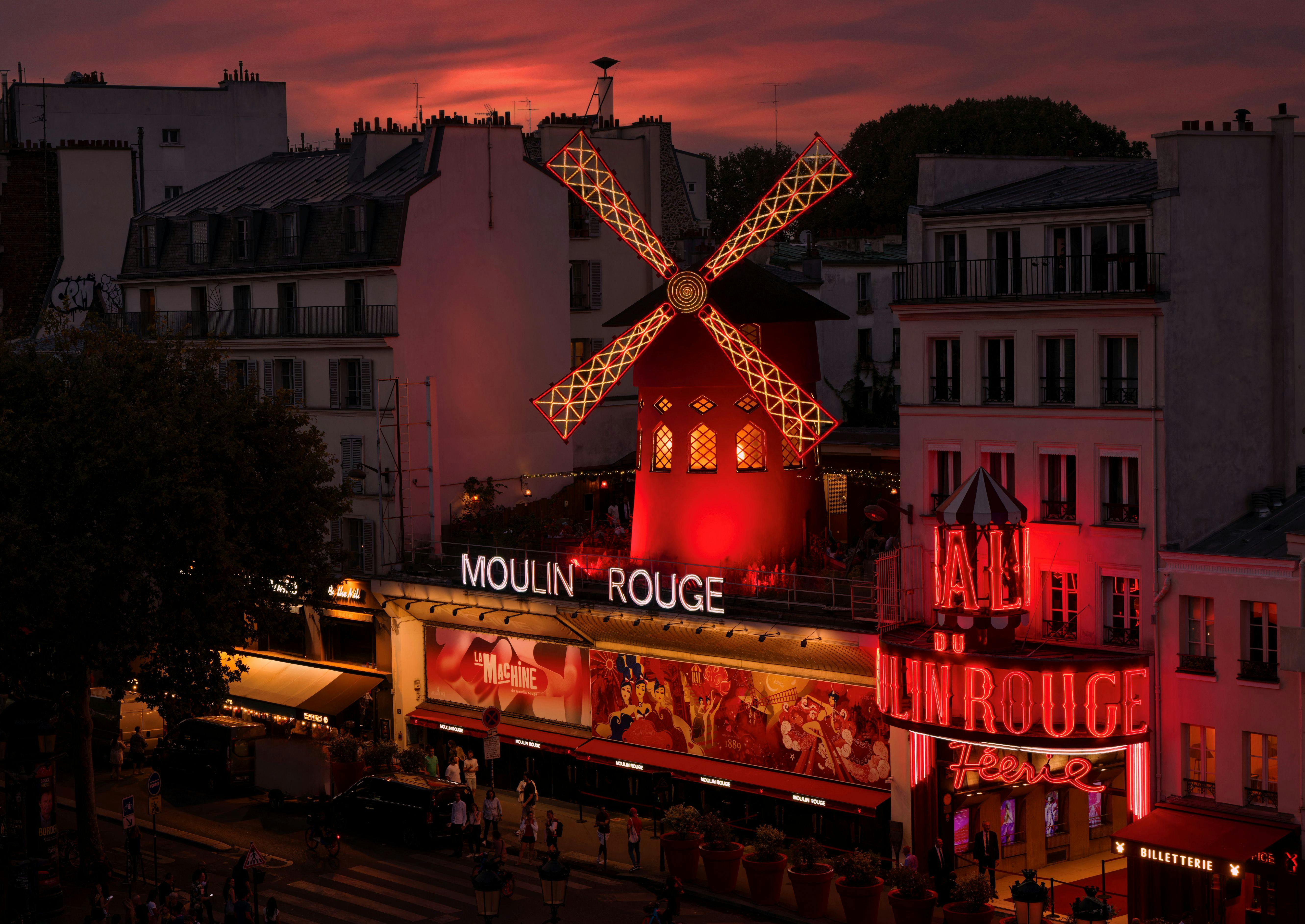 L'edifici il·luminat del Moulin Rouge amb el seu emblemàtic molí de vent vermell al capvespre, envoltat d'edificis de la ciutat i activitat al carrer.