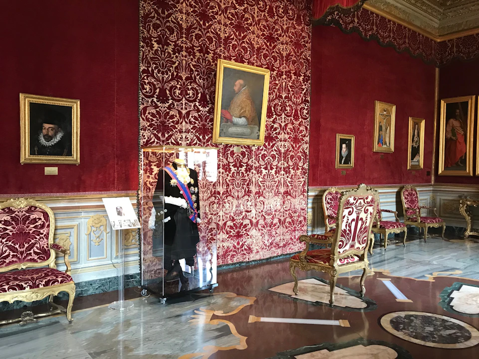 Ornate room with red damask walls, framed portraits, glass-encased uniform, and opulent chairs with gold detailing. Marble flooring.