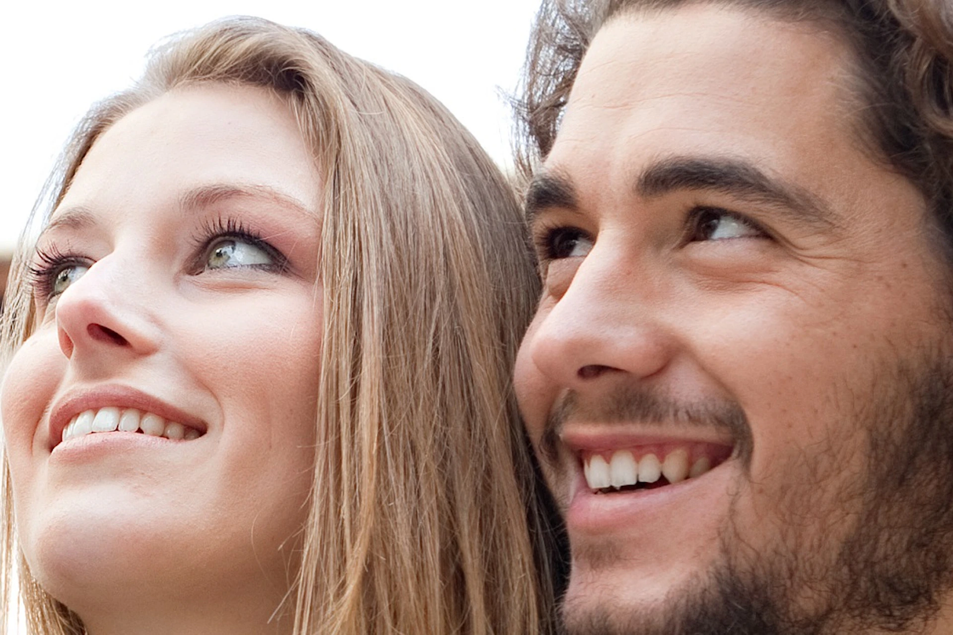 A smiling couple takes a selfie in an outdoor urban area, with buildings and a clear sky visible in the background.