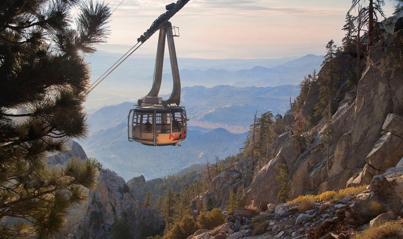 Aerial tramway car suspended above a rugged, mountainous landscape with trees and distant hazy mountains.