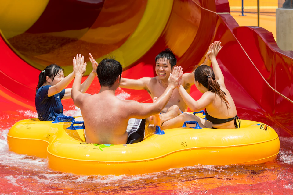 Three people in swimsuits on a yellow inflatable raft at a water park, high-fiving and smiling near a colorful water slide.