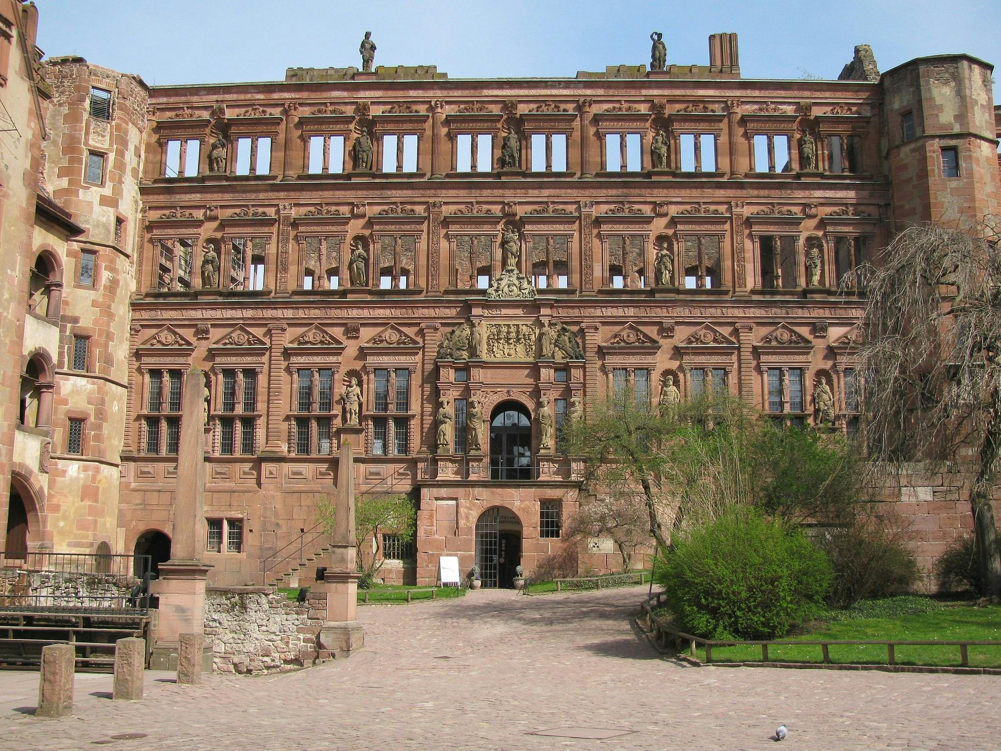 Historic red sandstone building with ornate facade, statues, archways, and a courtyard with greenery and cobblestones.