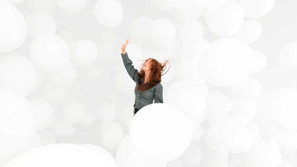 A person with curly hair wearing a striped shirt, surrounded by numerous large white balloons, reaching upward.
