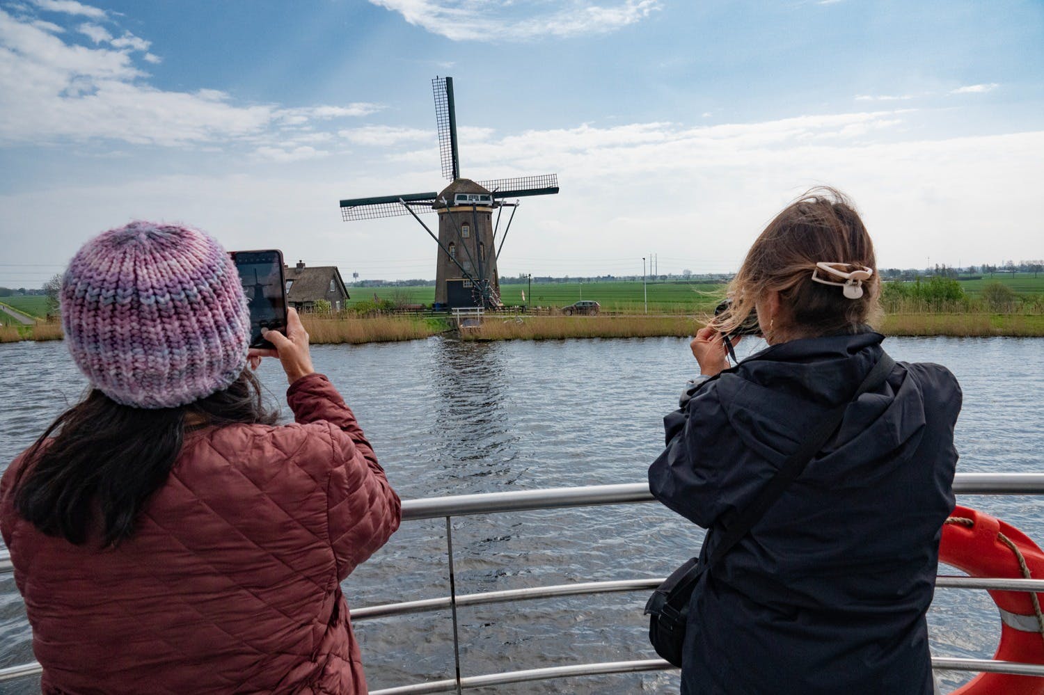 Two people take photos of a traditional windmill across a canal, with green fields and a cloudy sky in the background.