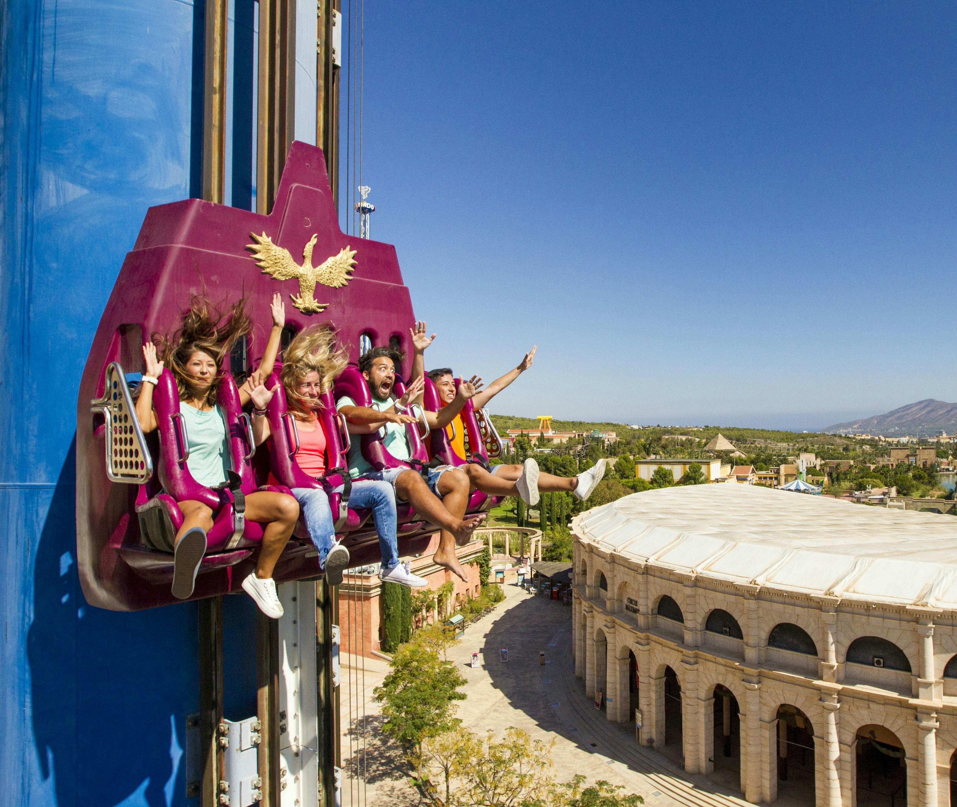 Persone su una giostra da brivido con i piedi a penzoloni e le mani in alto, sullo sfondo di un parco assolato e di un anfiteatro in stile romano.