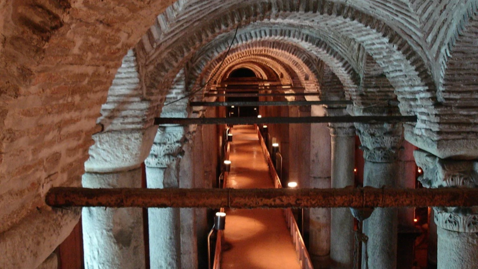 A dimly lit underground corridor with arched ceilings, stone columns, and a walkway illuminated by lights.