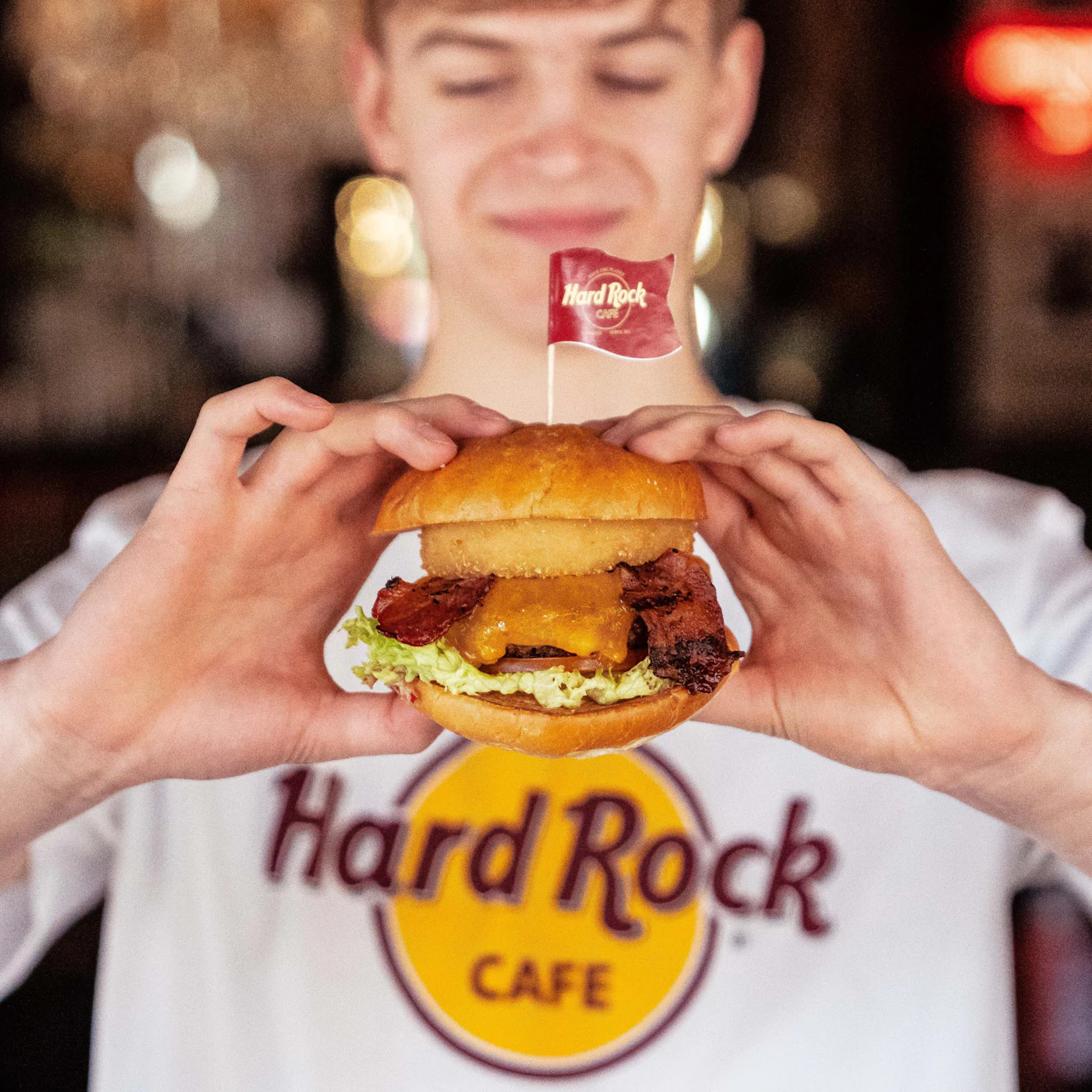 Person in a Hard Rock Cafe shirt holding a burger with a Hard Rock flag, featuring bacon, cheese, and lettuce.