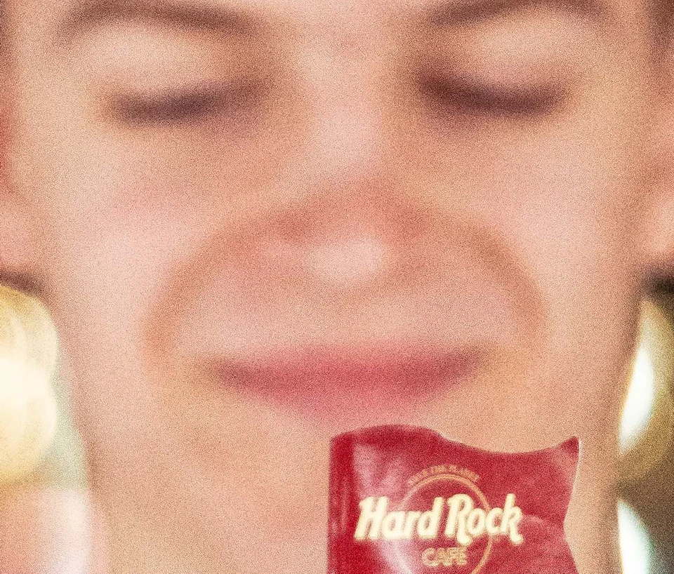 Person in a Hard Rock Cafe shirt holding a burger with a Hard Rock flag, featuring bacon, cheese, and lettuce.