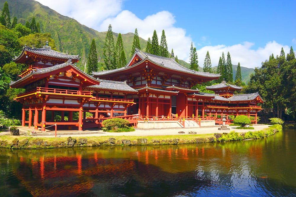 Red traditional Japanese building with ornate roof, located by a pond with reflections, surrounded by lush greenery and mountains.