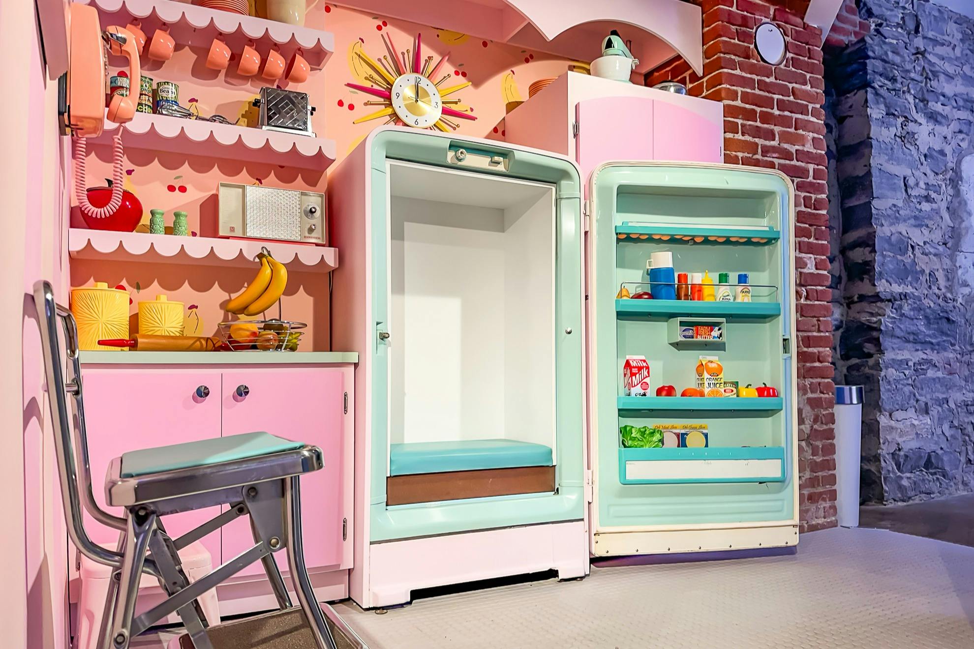 Μια ρετρό κουζίνα με ροζ θέμα, με ένα ανοιχτό vintage ψυγείο γεμάτο με φαγητό και έναν άδειο καταψύκτη.