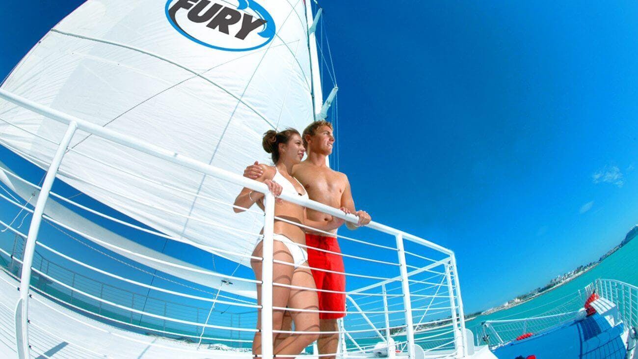 Una dona amb un banyador blanc i un home amb pantalons curts vermells s'aixequen en un veler amb una vela etiquetada "Fury" contra un cel blau clar.