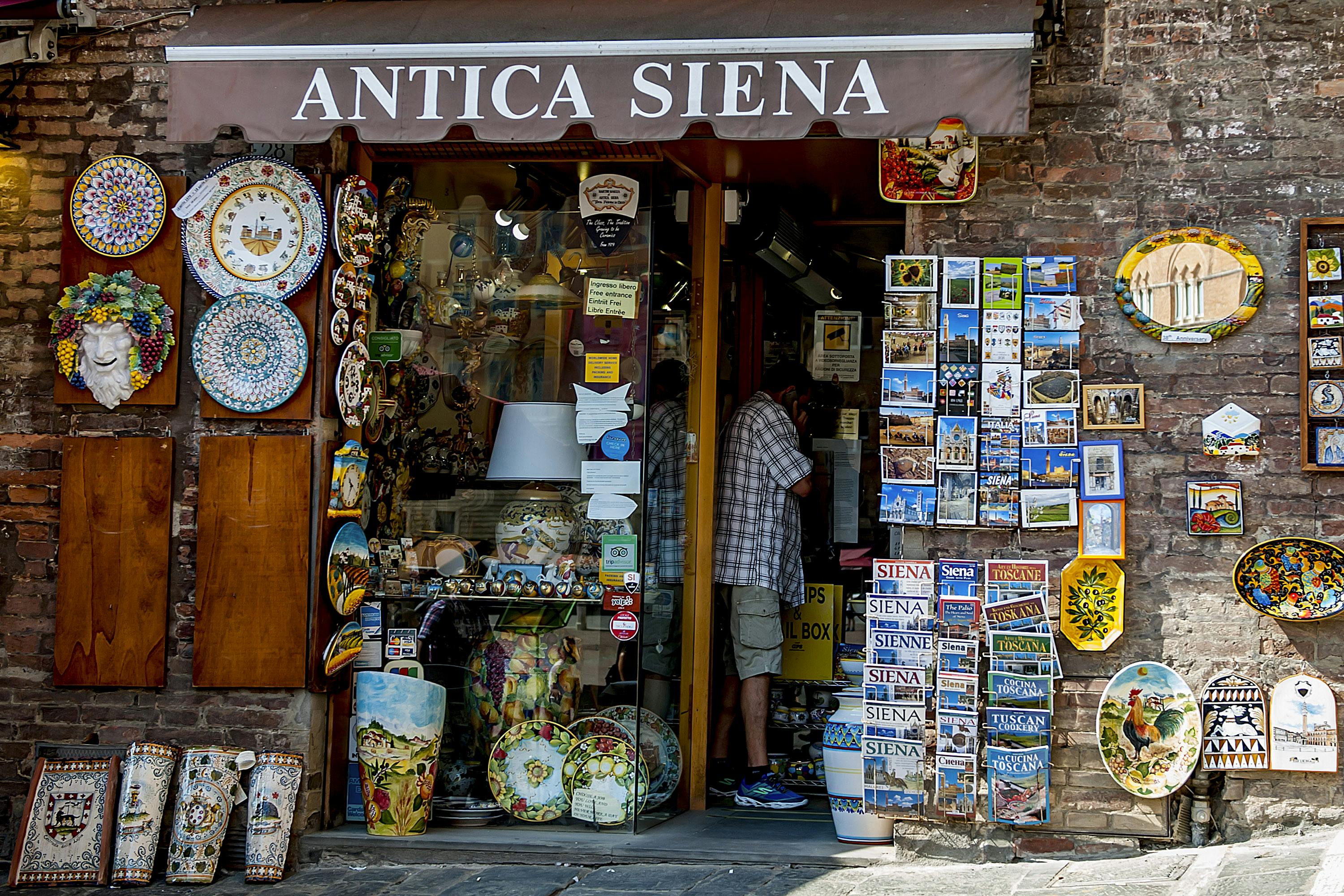 一个标有“Antica Siena”的繁忙店面，展示陶瓷、明信片和纪念品，里面有人在浏览。