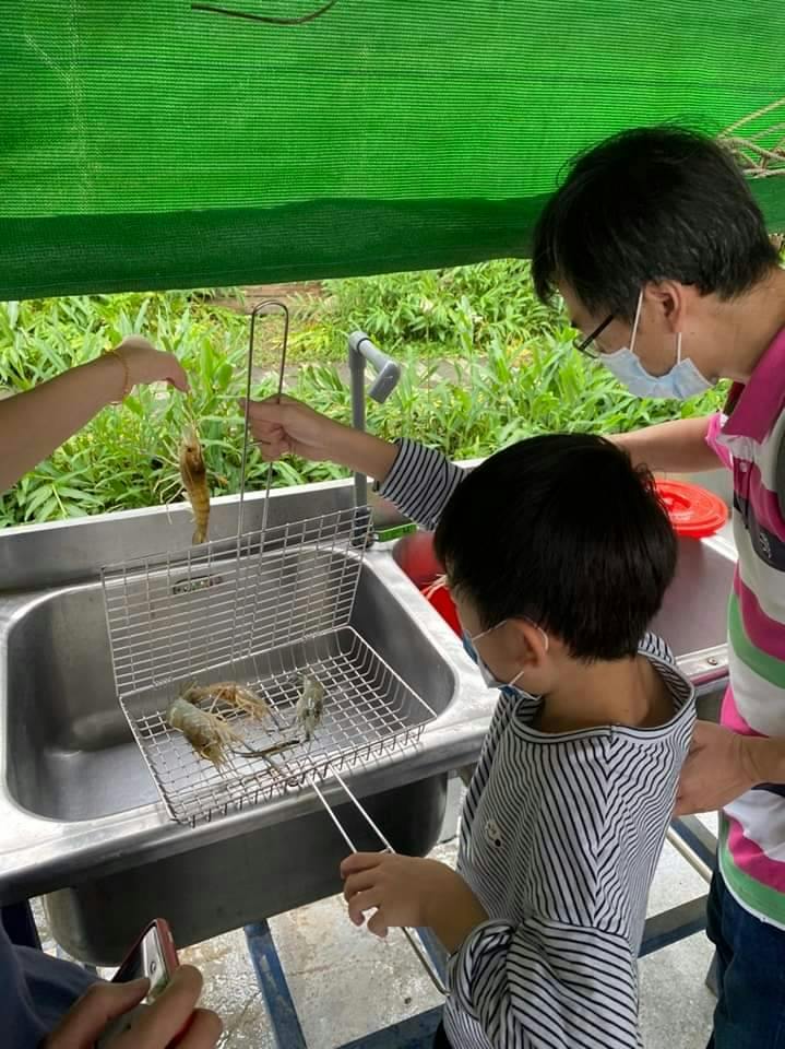 A child and an adult, both wearing masks, examining a basket containing prawns at a sink under a green canopy.