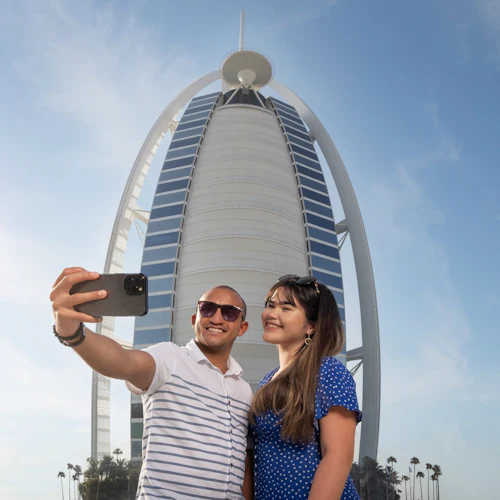 A couple takes a selfie in front of the Burj Al Arab building under a clear blue sky.