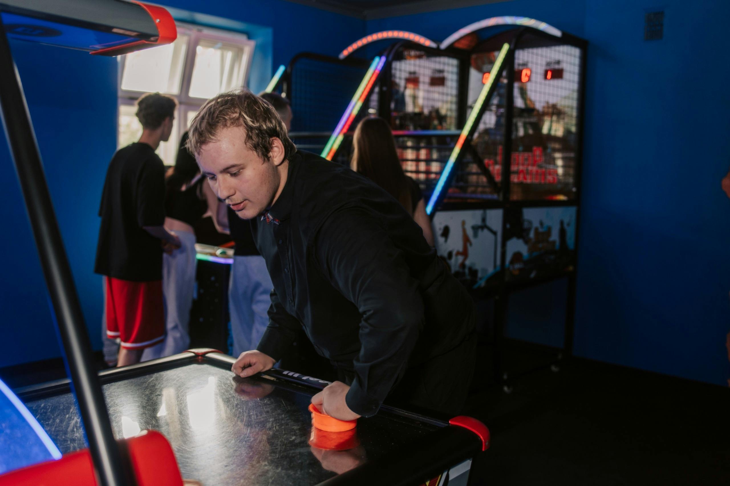 A person playing air hockey with a red paddle in an arcade, with other people and arcade machines in the background.