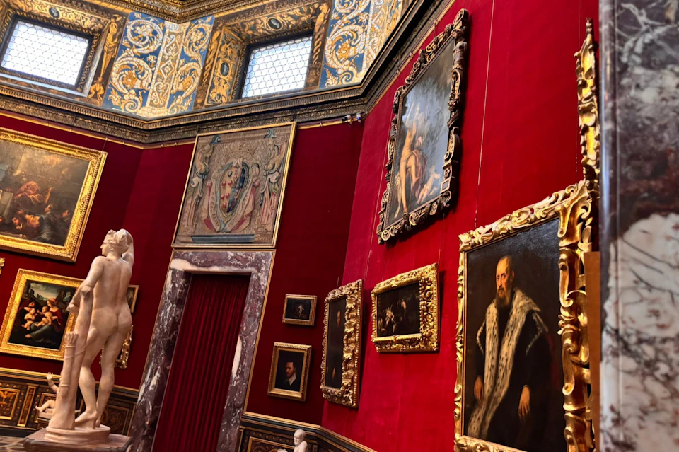 An ornate gallery with red walls, gilded picture frames, classical paintings, and marble sculptures on display.