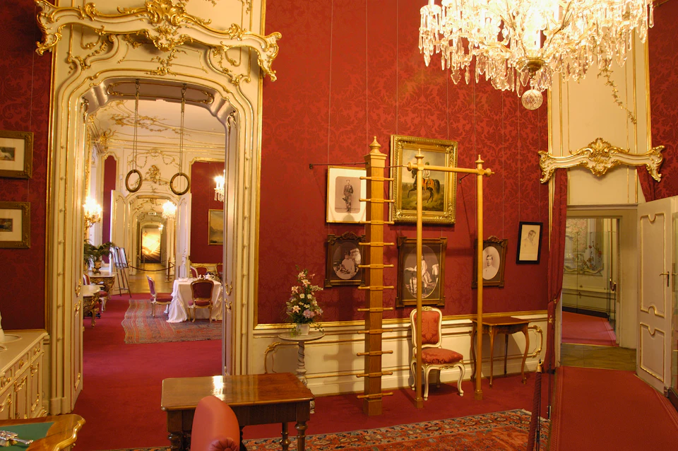 Ornate room with red walls, gold accents, chandelier, framed portraits, chairs, and tables. Doorway leads to another similarly decorated room.