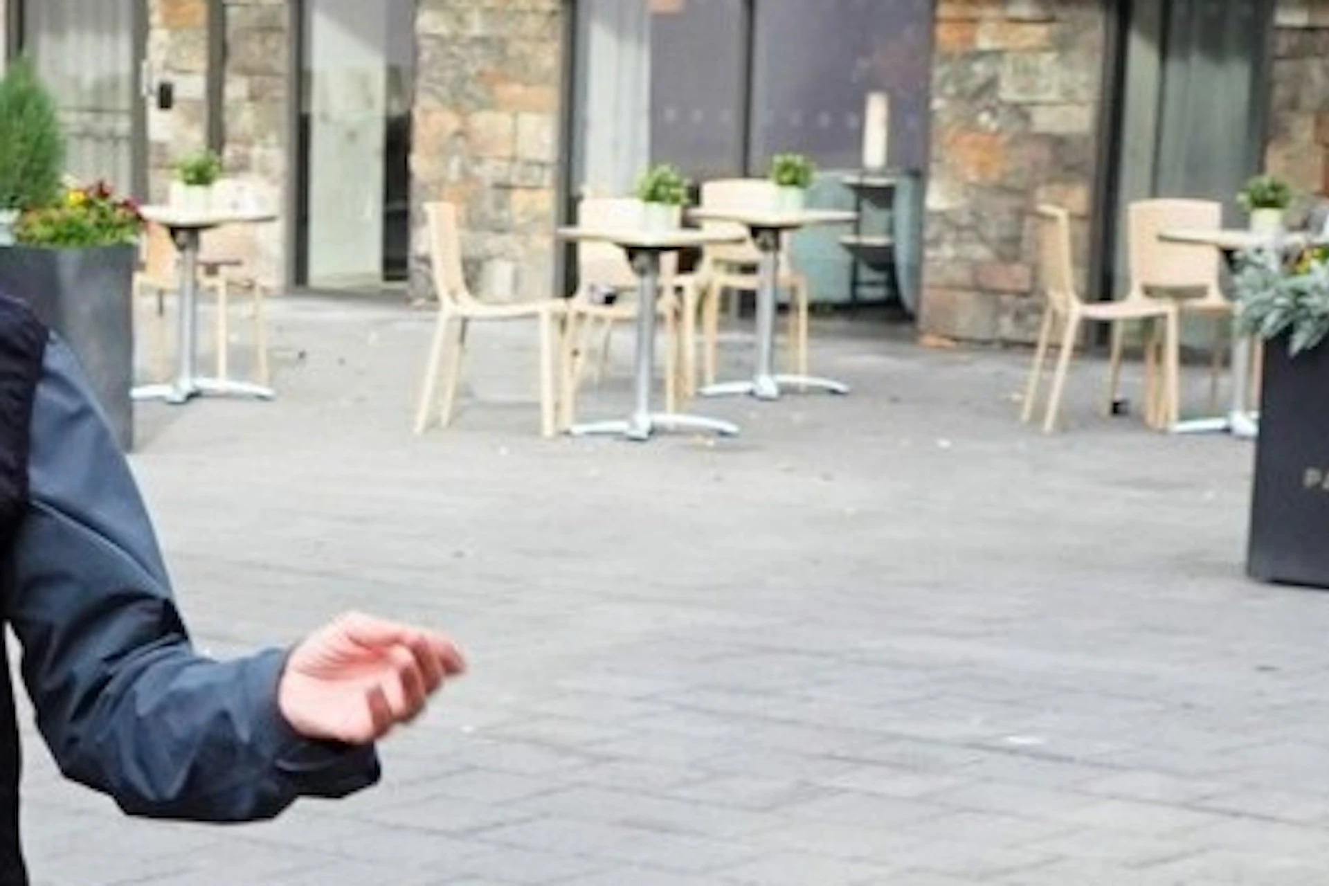 A man in outdoor clothing gesturing while speaking to a group of people in a paved courtyard area near a stone building.