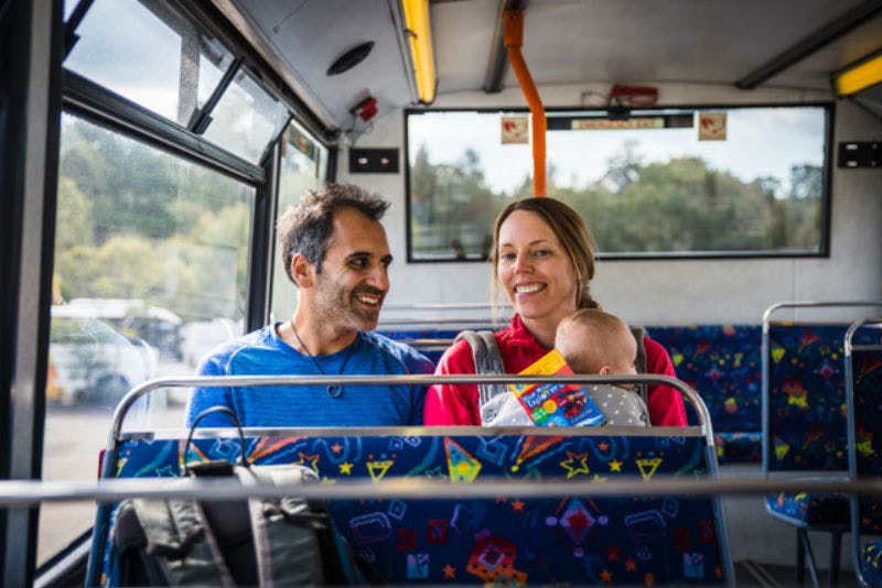 A smiling couple sits on a bus, the woman holding a baby who is looking at a colorful book. The bus seats have playful patterns.