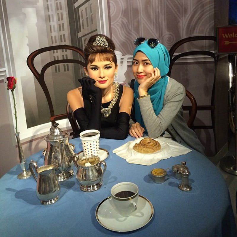 파란색 머리 스카프를 두른 한 여성이 조식과 식기가 놓인 테이블에서 오드리 헵번의 왁스 동상을 들고 포즈를 취하고 있습니다.