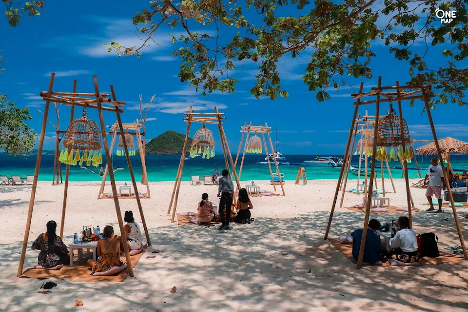 People relaxing on a sandy beach under shaded wooden structures with hanging decorations, clear blue water, and boats in the background.
