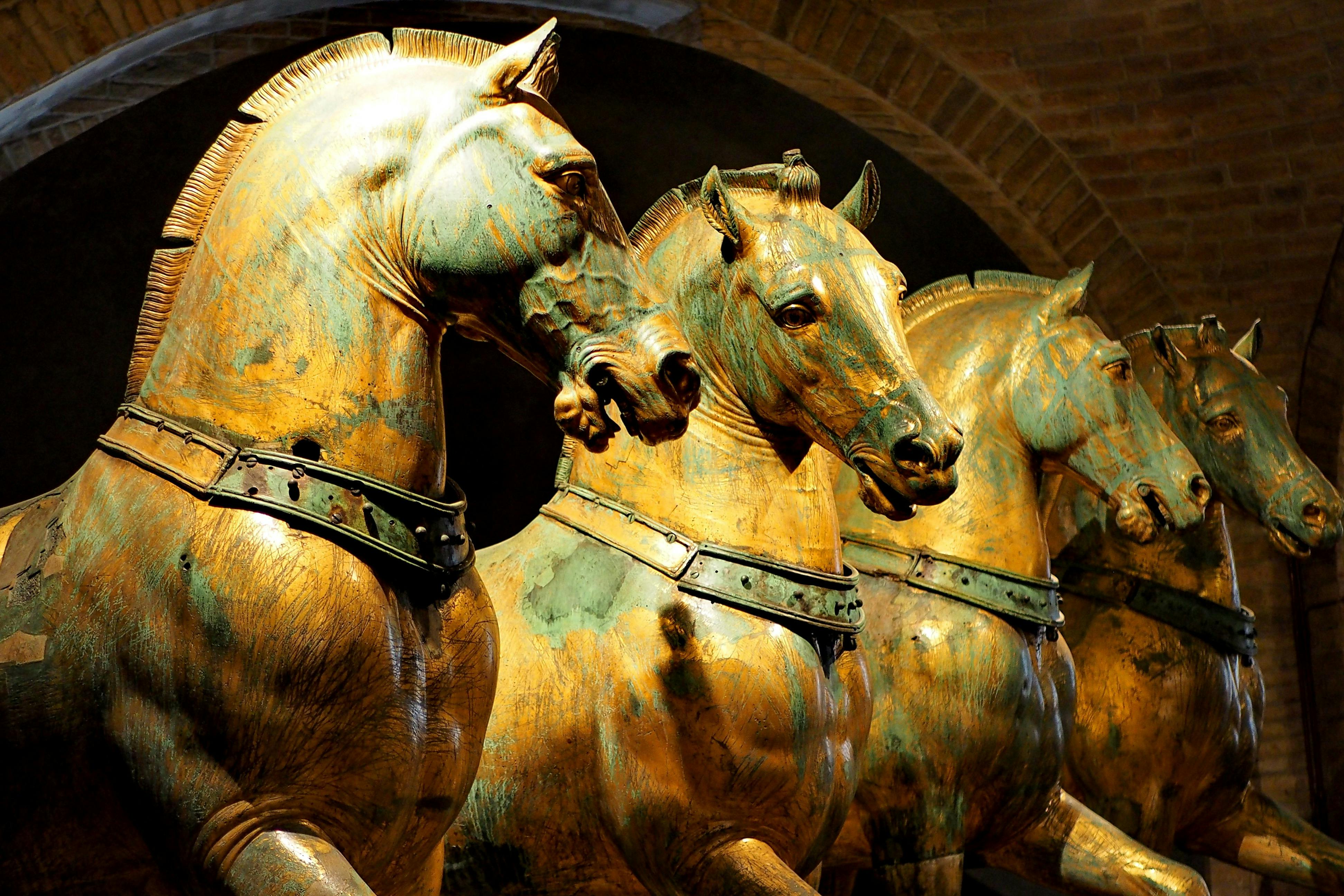 Three golden horse sculptures with green patina, wearing harnesses, displayed in an arched indoor setting.
