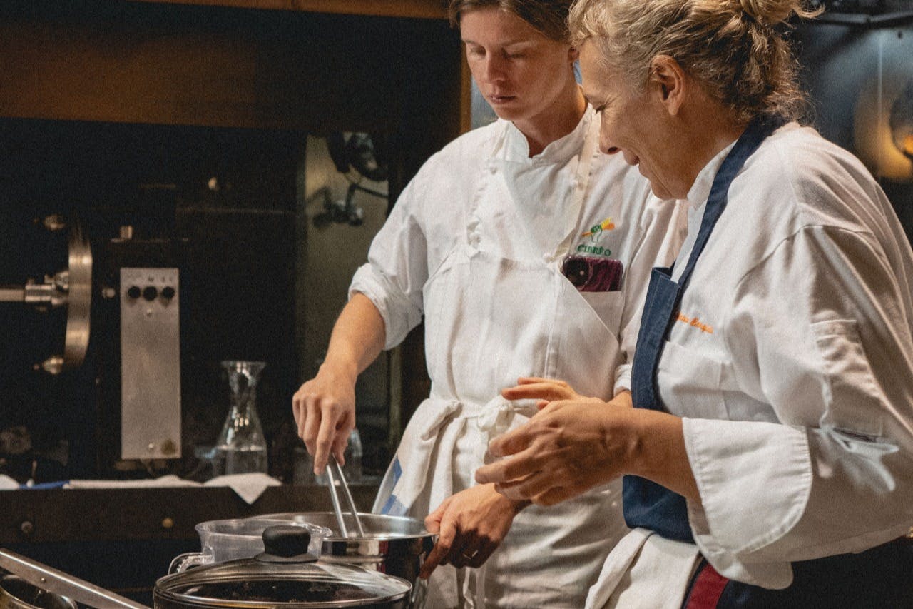 Two chefs in white uniforms cooking, one stirring a pot while the other observes.