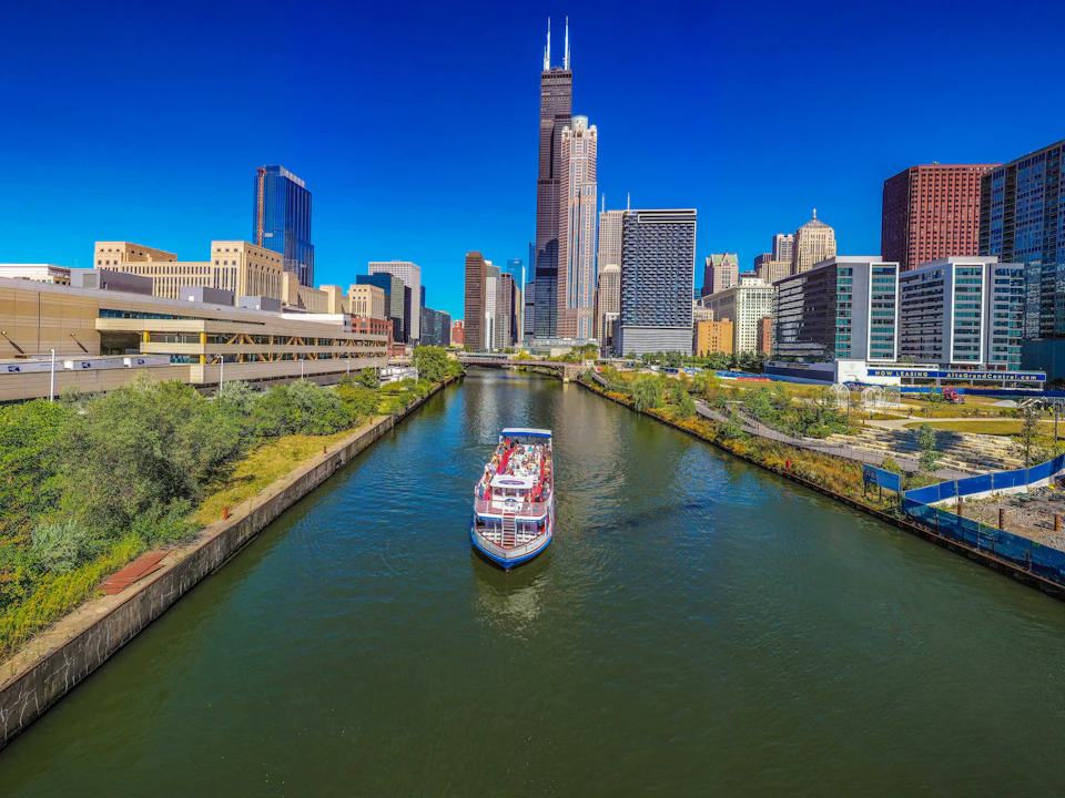 A boat cruises along a city river with tall skyscrapers, including a distinctive two-pronged building, under a clear blue sky.
