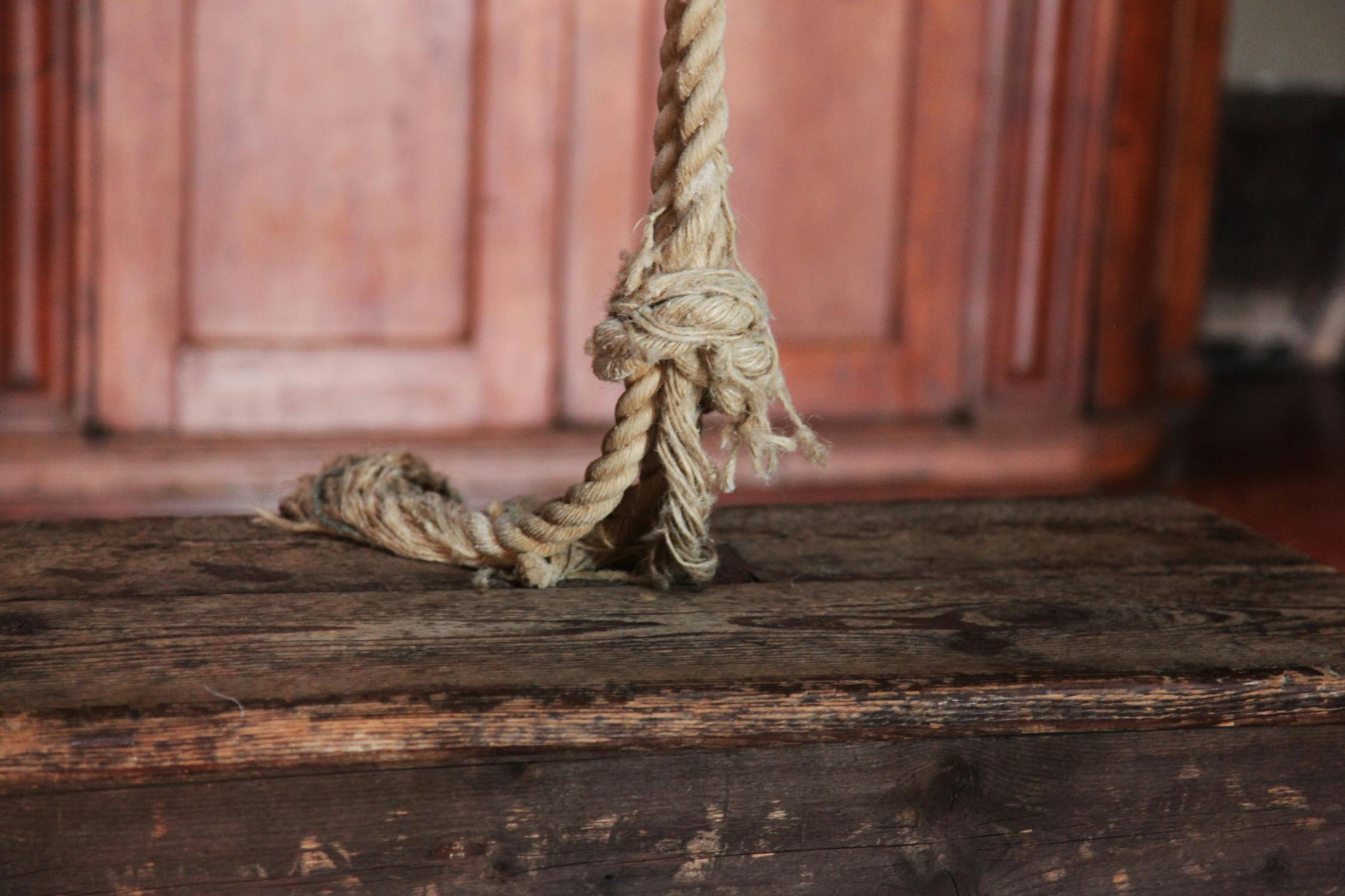 Κοντινό πλάνο ενός ξεφτισμένου σχοινιού δεμένου γύρω από ένα ξύλινο δοκάρι με μια θολή ξύλινη πόρτα στο παρασκήνιο.