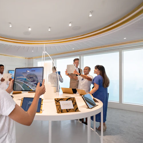People in a modern room using tablets, surrounded by display tables with models and screens. White and gold ornate ceiling.