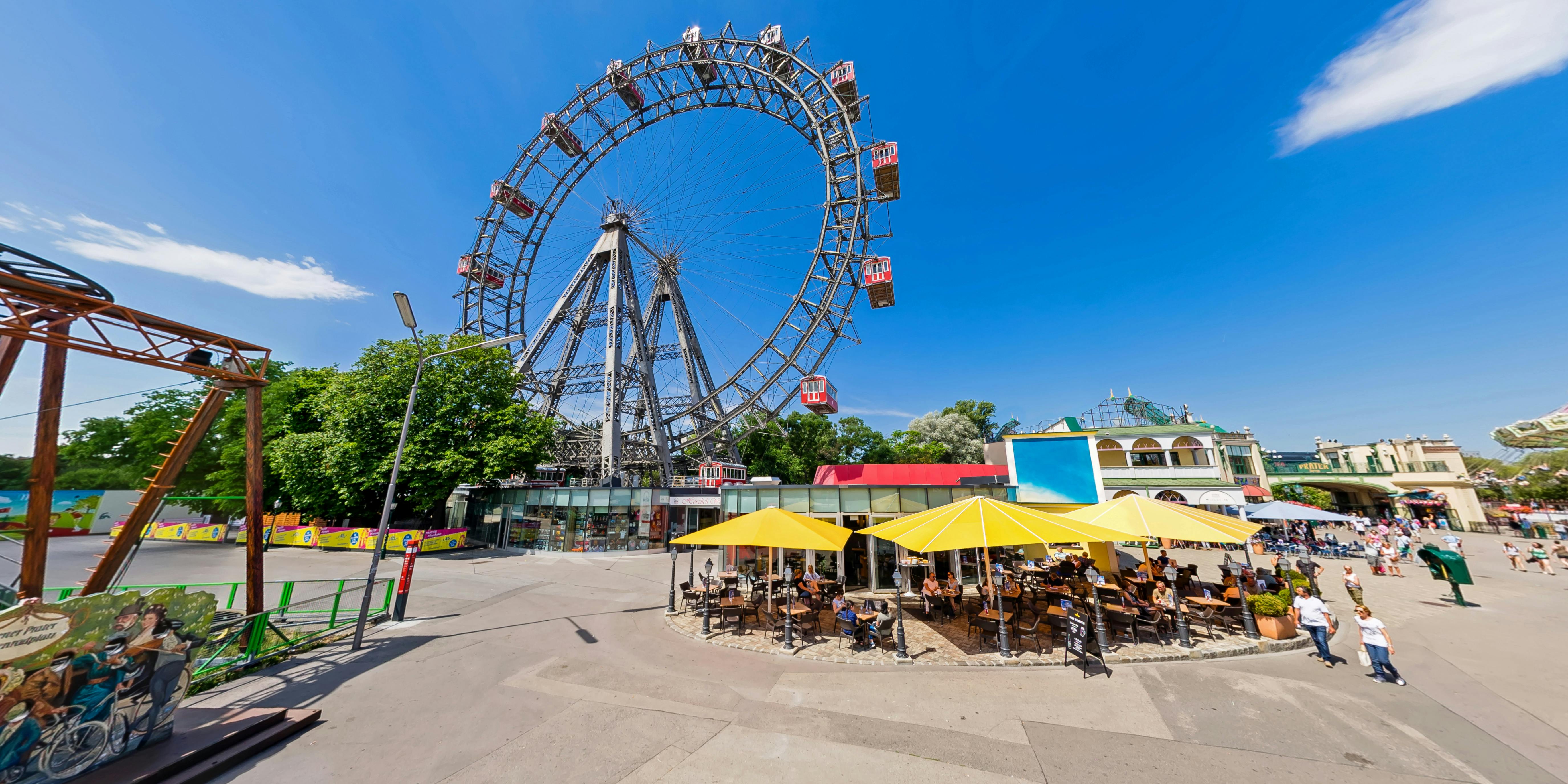 Nad kavárnou se žlutými deštníky se tyčí ruské kolo s červenými osobními vozy. Lidé sedí u venkovních stolů.