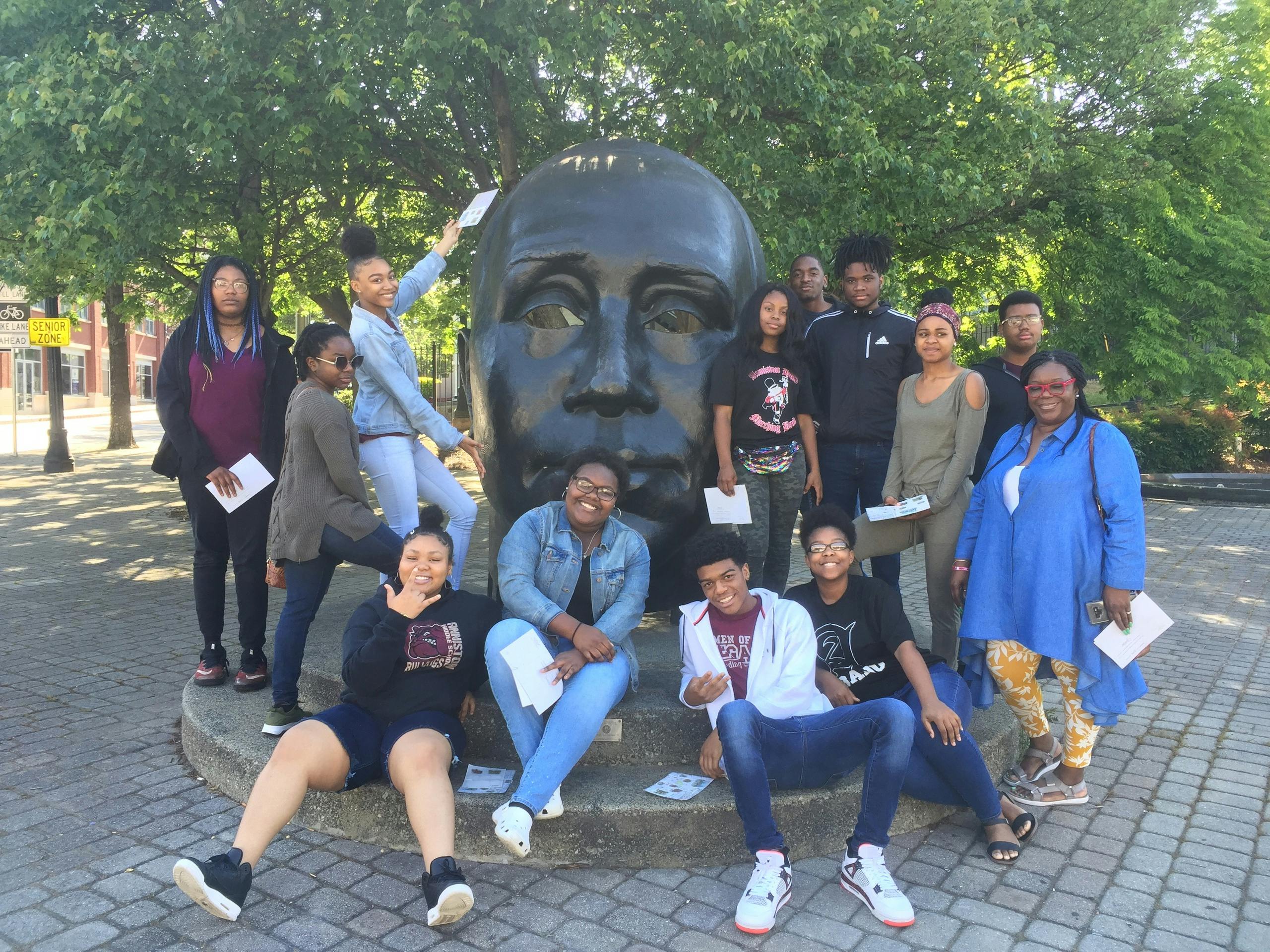 Un groupe de personnes posant devant une grande statue de tête dans un parc avec des arbres en arrière-plan.