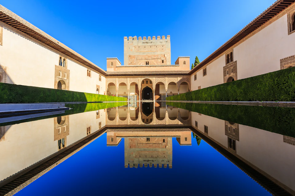 Symmetrical courtyard with archways, reflected perfectly in a long, rectangular pool under a clear blue sky.