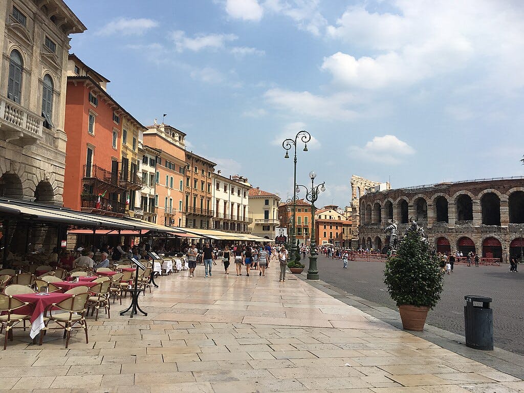 Venkovní kavárna se stoly a červenými ubrusy, procházky lidí, barevné budovy a starobylý amfiteátr pod modrou oblohou.