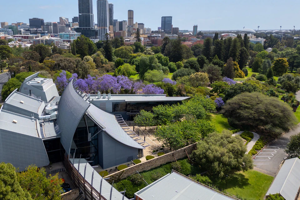Aerial view of a modern building surrounded by greenery and purple flowering trees, with a city skyline in the background.