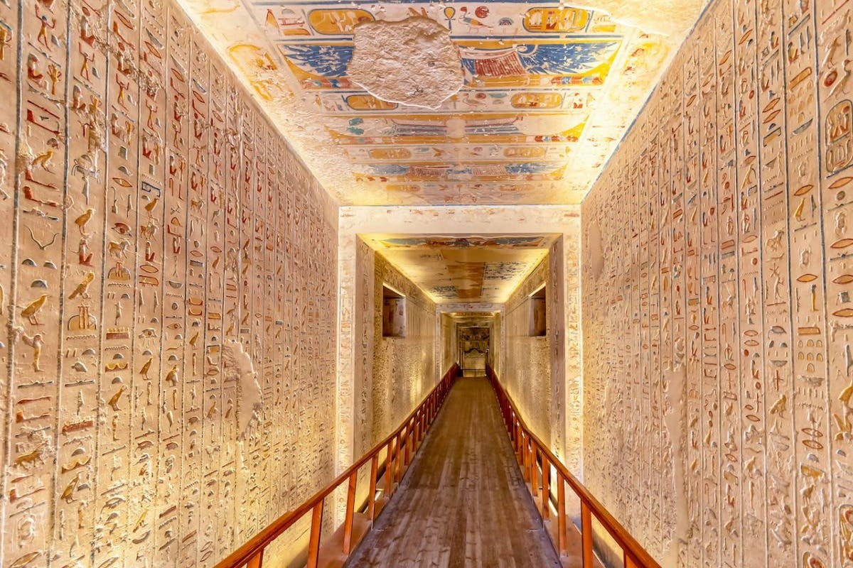 A long, narrow, ancient Egyptian corridor with walls and ceiling covered in hieroglyphics and colorful paintings. Wooden railings line the walkway.