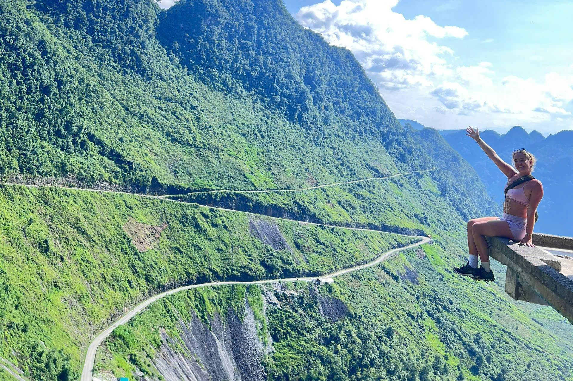 En person, der sidder på en klippekant med panoramaudsigt over en frodig, grøn bjergskråning og snoede veje under en lys himmel.