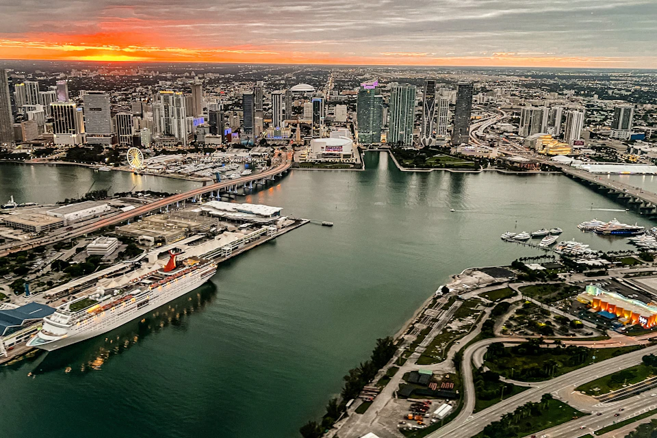 Aerial view of a cityscape at sunset with high-rise buildings, a bridge, a docked cruise ship, and a waterfront area.