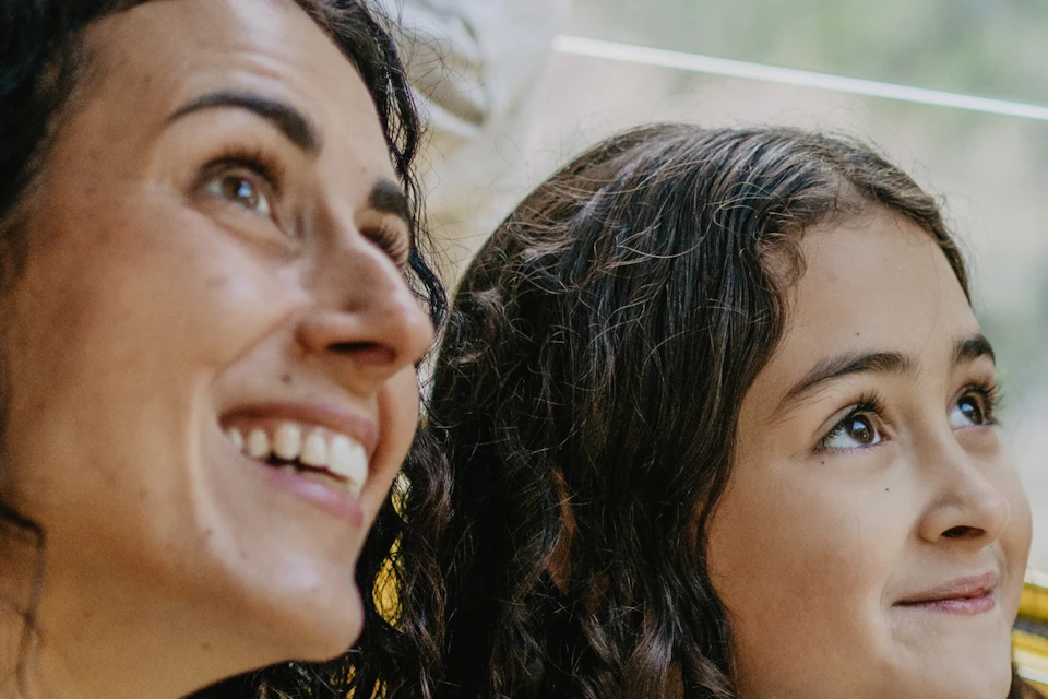 A woman and a young girl smiling while looking out of a window, with blurred greenery visible outside.