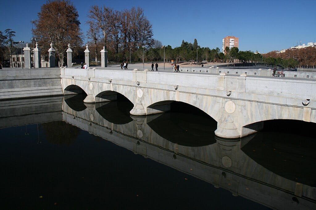 A stone bridge with multiple arches over calm water, with people walking and cycling on it, surrounded by trees and a clear sky.