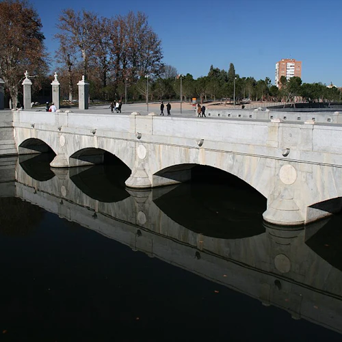 En stenbro med flera valv över lugnt vatten, med människor som går och cyklar på den, omgiven av träd och en klar himmel.