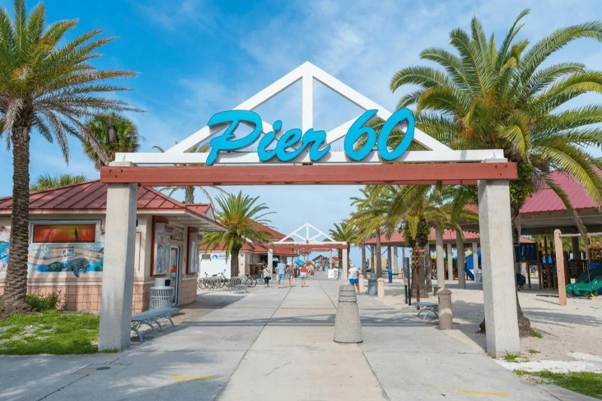 Entrance to Pier 60 with a blue sign, palm trees, and a walkway leading to the beach under a blue sky.